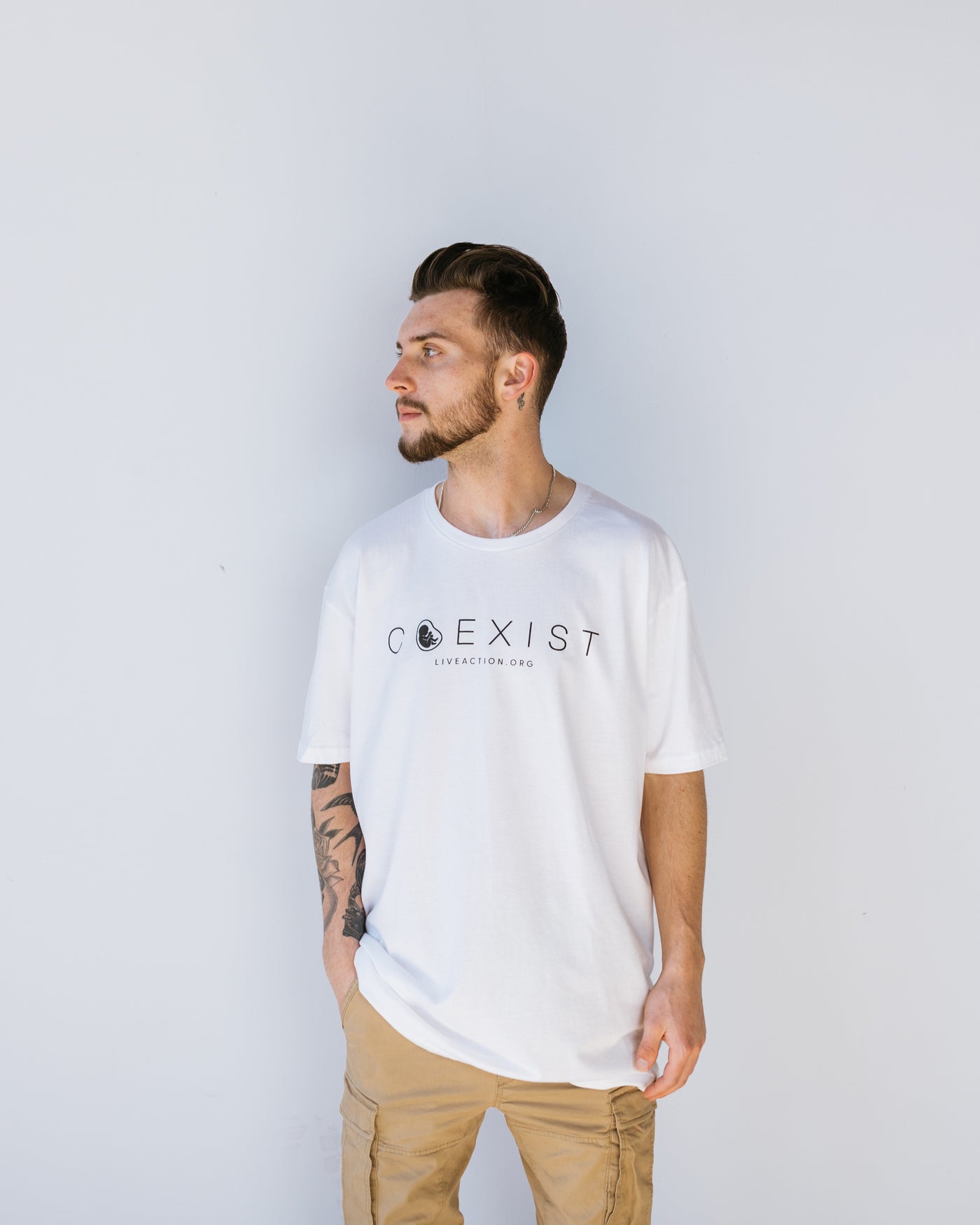 A man with brown hair and a short beard in a white tee with the word "COEXIST" written across the chest in fine black font. Inside the misshapen O is a preborn baby. Underneath the main text is Live Action's website written in small font.