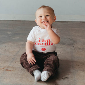Cute baby smiling in a shirt that says "i am pro-life", brown corduroy pants, and grey striped socks.