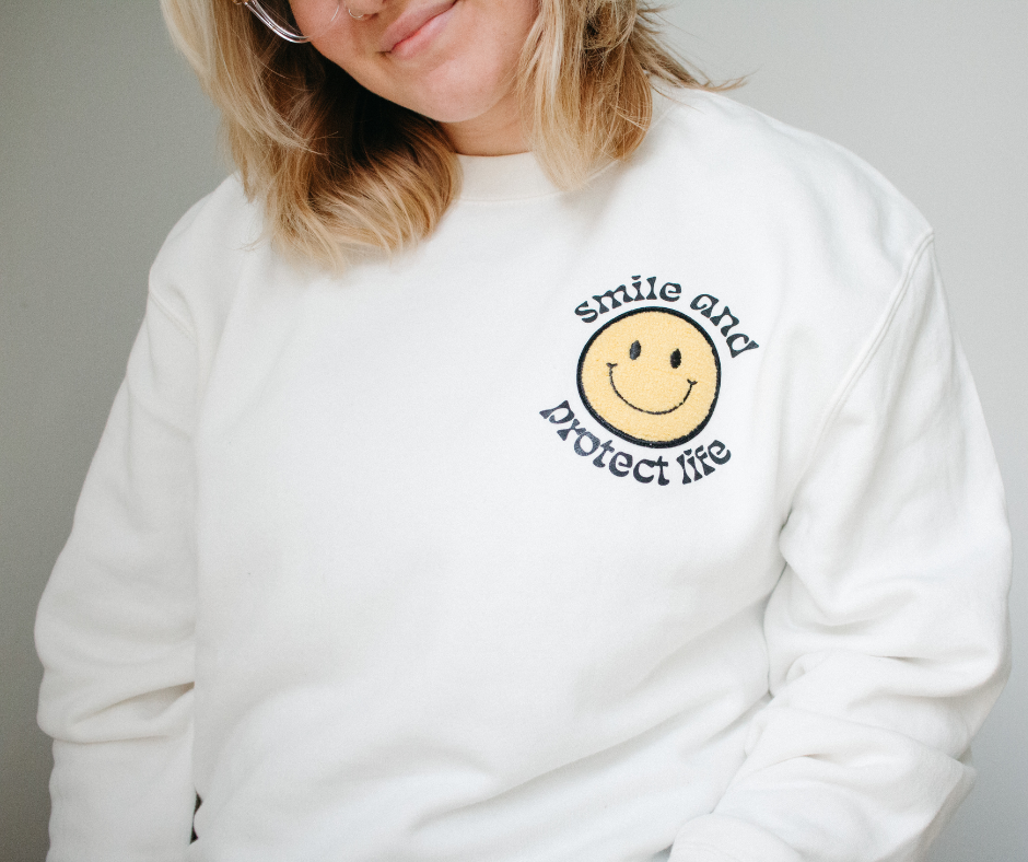 A blonde woman in a bone white, crewneck sweatshirt with a smiley patch in the upper right hand corner of the shirt, surrounded by the words "smile and protect life."