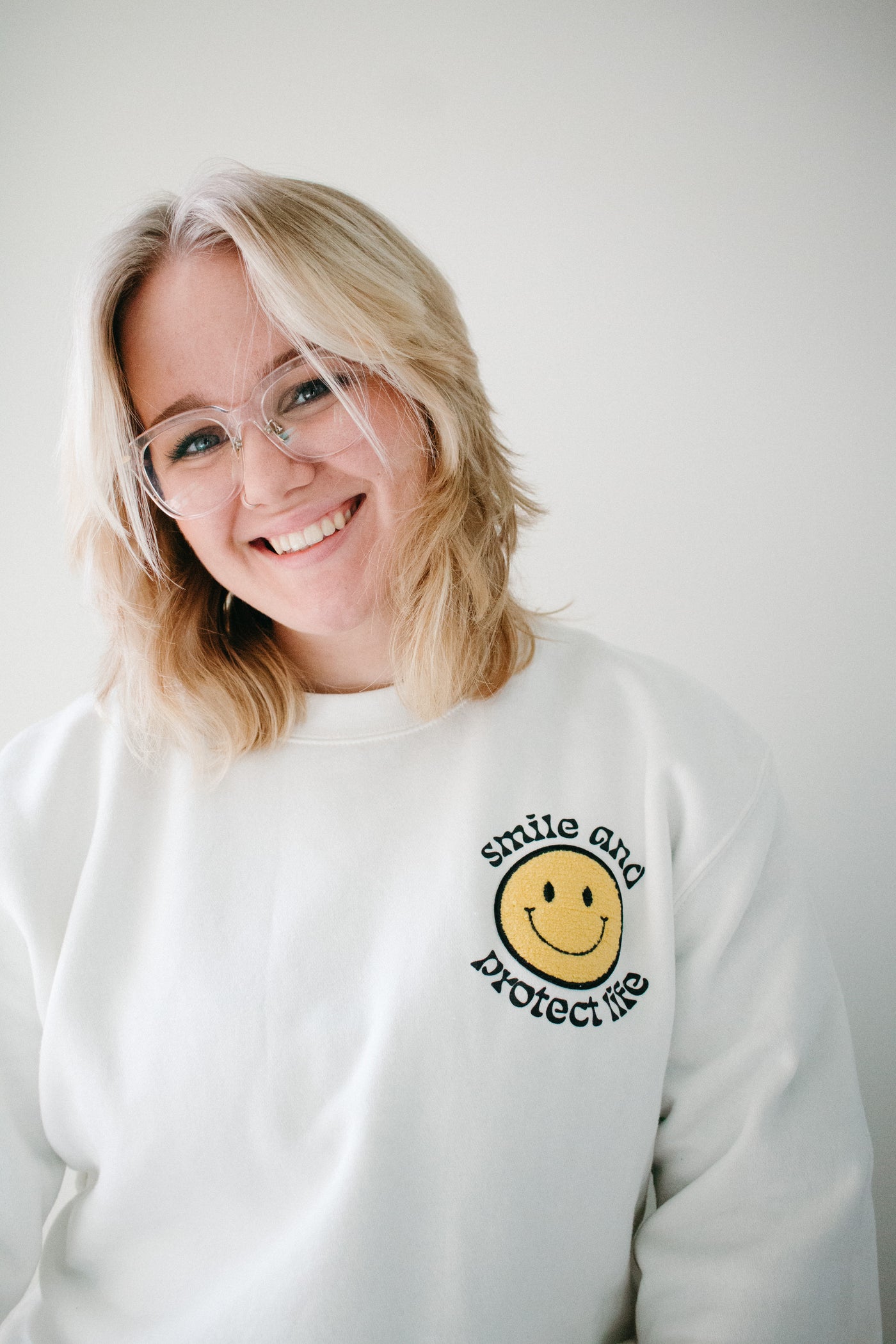 A blonde woman in a bone white, crewneck sweatshirt with a smiley patch in the upper right hand corner of the shirt, surrounded by the words "smile and protect life."