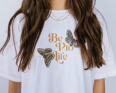 Closeup of a brunette woman in a white tee shirt with the words "Be Pro Life" in the center surrounded by two large green and brown butterflies.