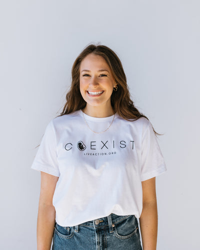 A woman with brown hair in a white tee with the word "COEXIST" written across the chest in fine black font. Inside the misshapen O is a preborn baby. Underneath the main text is Live Action's website written in small font.