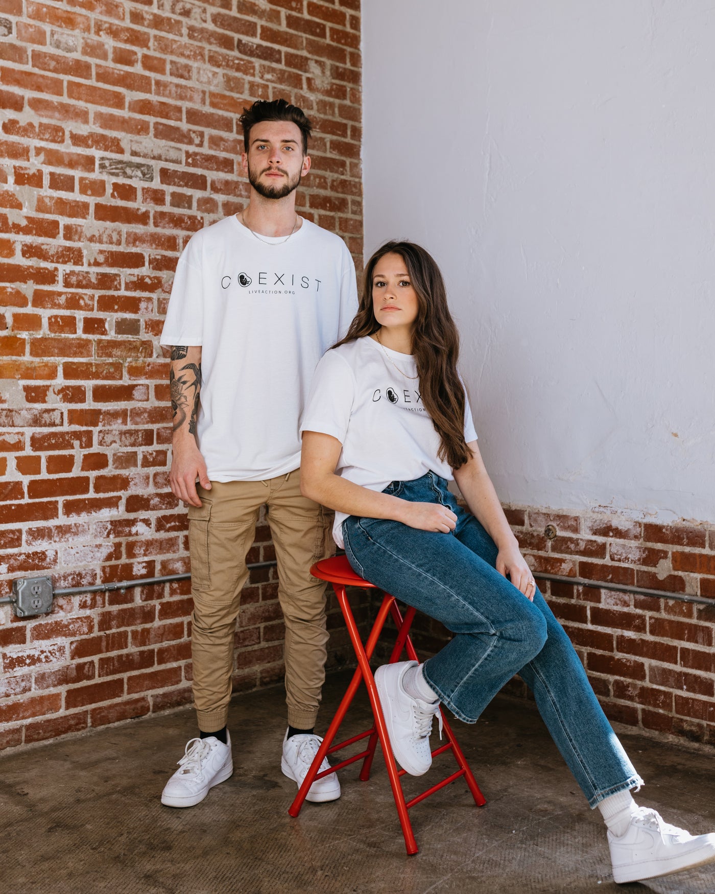 A man and woman both in white tees with the word "COEXIST" written across the chest in fine black font. Inside the misshapen O is a preborn baby. Underneath the main text is Live Action's website written in small font. 